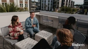 Four people chat over coffee on a sunny apartment rooftop terrace with mesh chairs and white cube seats, surrounded by apartment buildings, with the foshee logo at lower right.